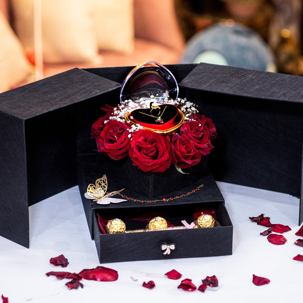Black jewelry box with red roses and gold jewelry on a white surface with rose petals.