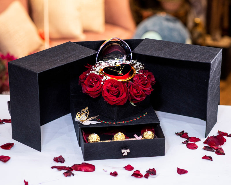 Black jewelry box with red roses and gold jewelry on a white surface with rose petals.