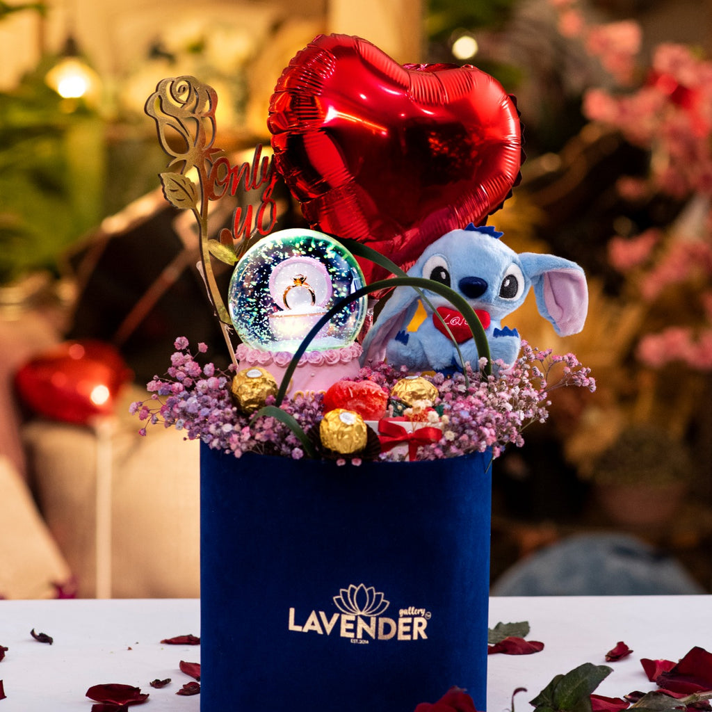 Decorative flower arrangement with a blue container, plush toy, and balloons on a table with rose petals.