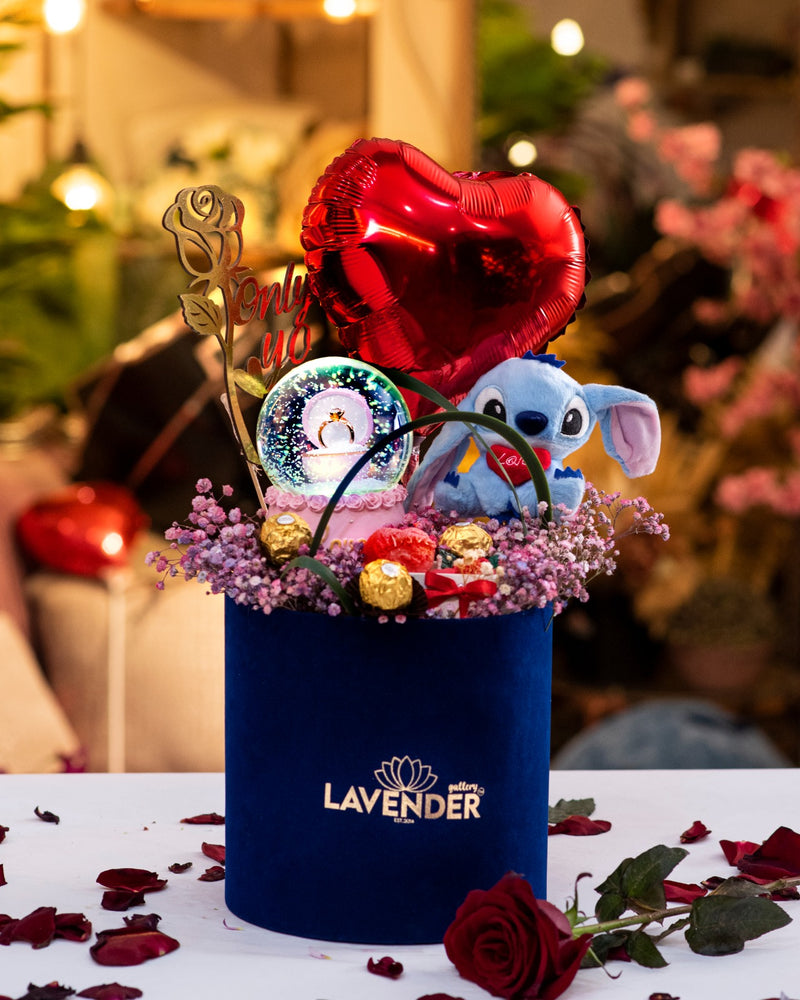 Decorative flower arrangement with a blue container, plush toy, and balloons on a table with rose petals.